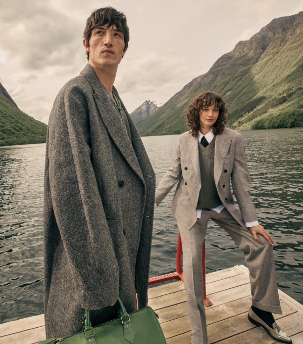 A man and woman wearing smart tailored suits standing on a jetty by a lake. 