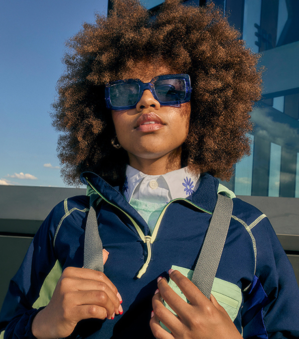 Woman wearing blue sunglasses and a navy jacket, holding backpack straps near a glass building.
