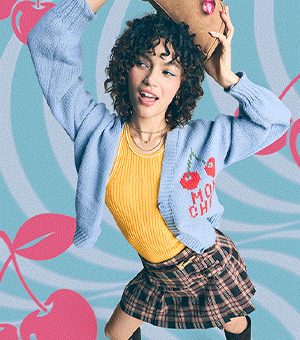 A woman, wearing a yellow vest, pale blue cardigan with cherry detailing and a plaid skirt, holds a tan bag over her head.